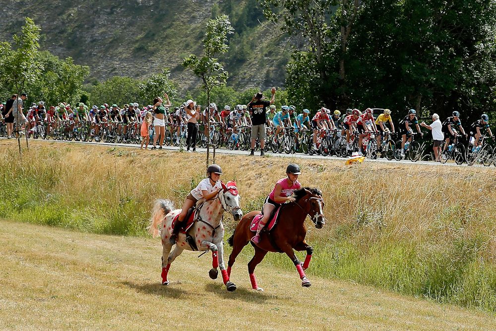 A pair of horses and riders gallop alongside the peloton during stage 17.