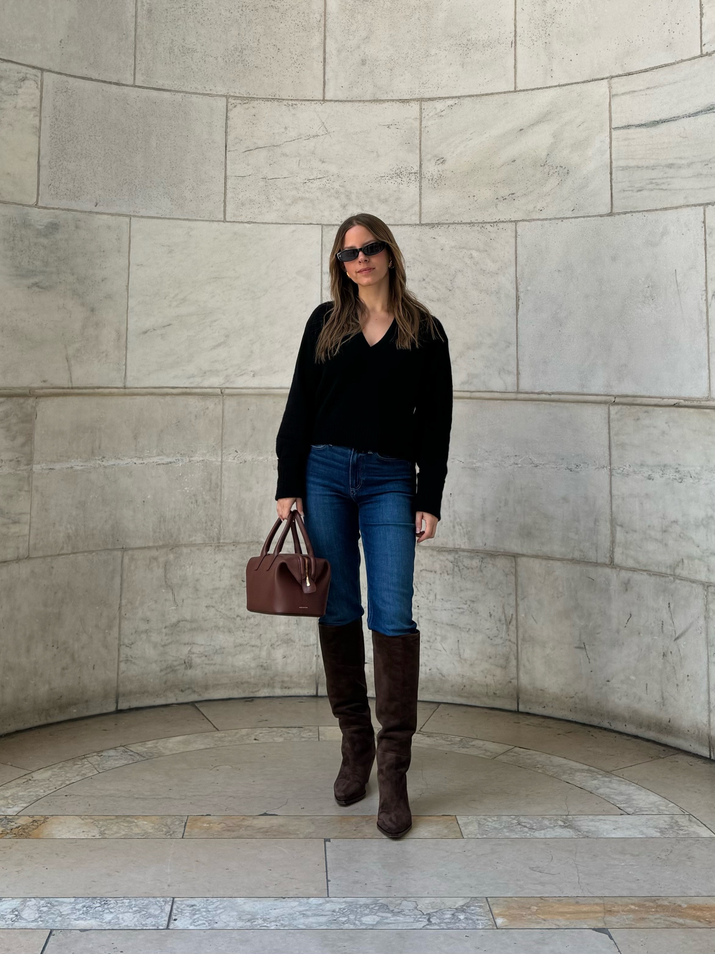 Nikki wears a black v-neck sweater, blue jeans, brown suede boots, and a brown bag.