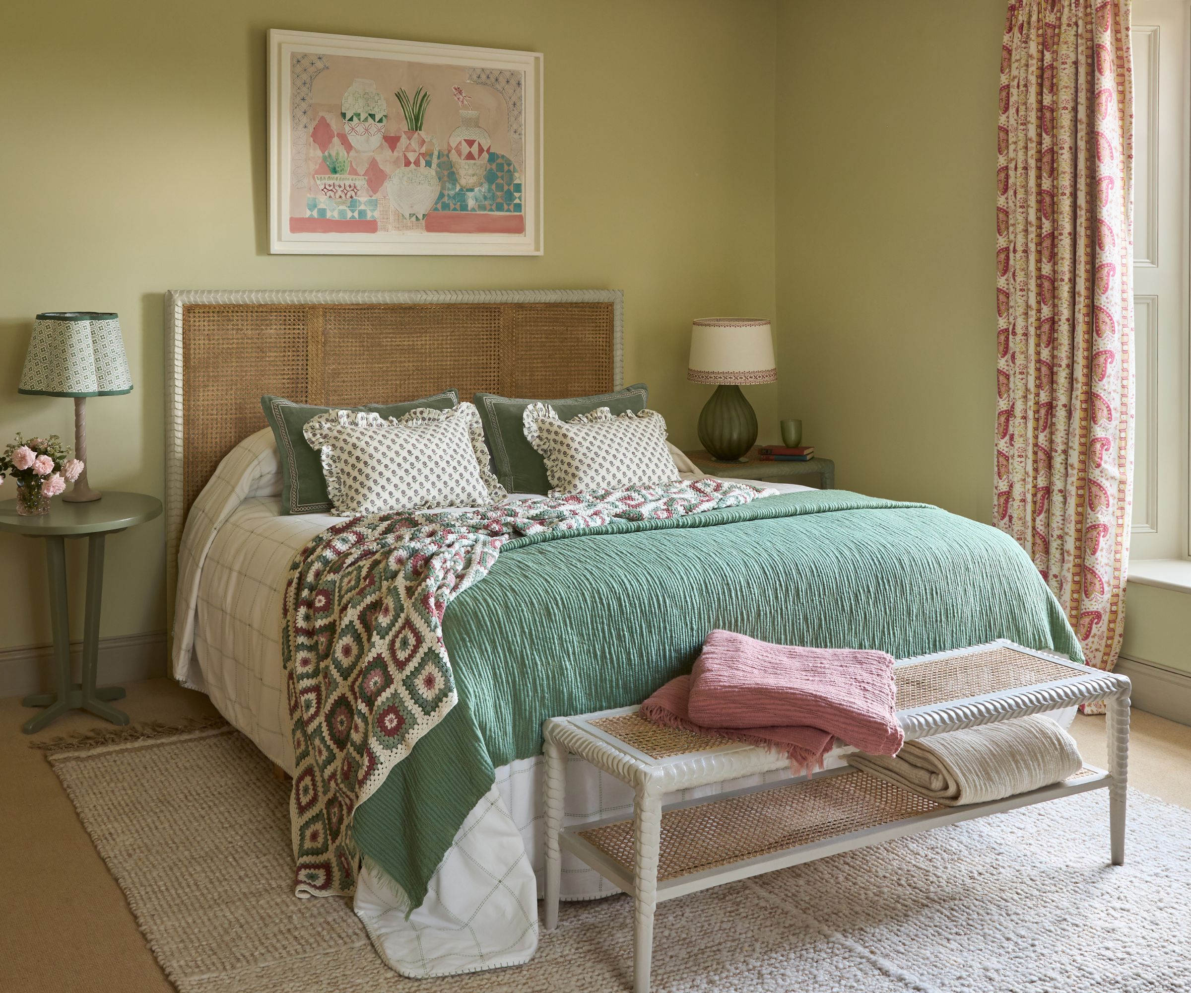 soft pale green bedroom styled with a rattan headboard, green bedspread with a crochet throw draped over the end. Pink paisley floral curtains frame the window