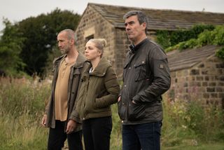 Grieving Sam, Belle and Cain stand outside and look off into the distance in Emmerdale 