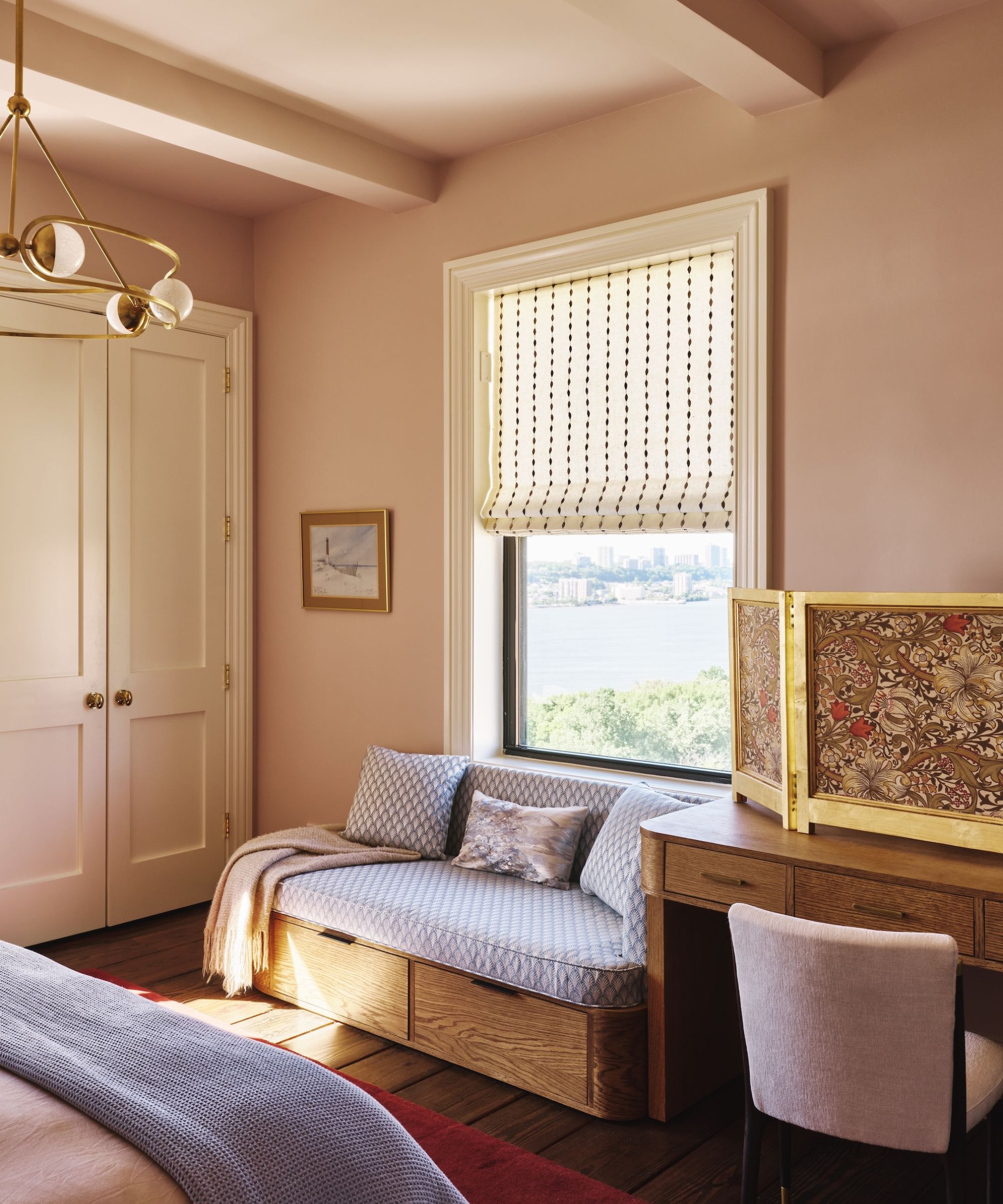 A calming bedroom with soft pink walls, a built-in wardrobe, a window with a cream patterned blind, a vanity table with a chair, and a window seat upholstered with blue patterned fabric