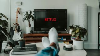 A person sits with their feet up on a coffee table. In the background the Netflix logo is displayed on a TV
