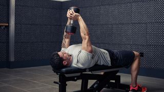Man in starting position for dumbbell pull-over exercise, lying on a flat weights bench with one dumbbell held above his head in both hands
