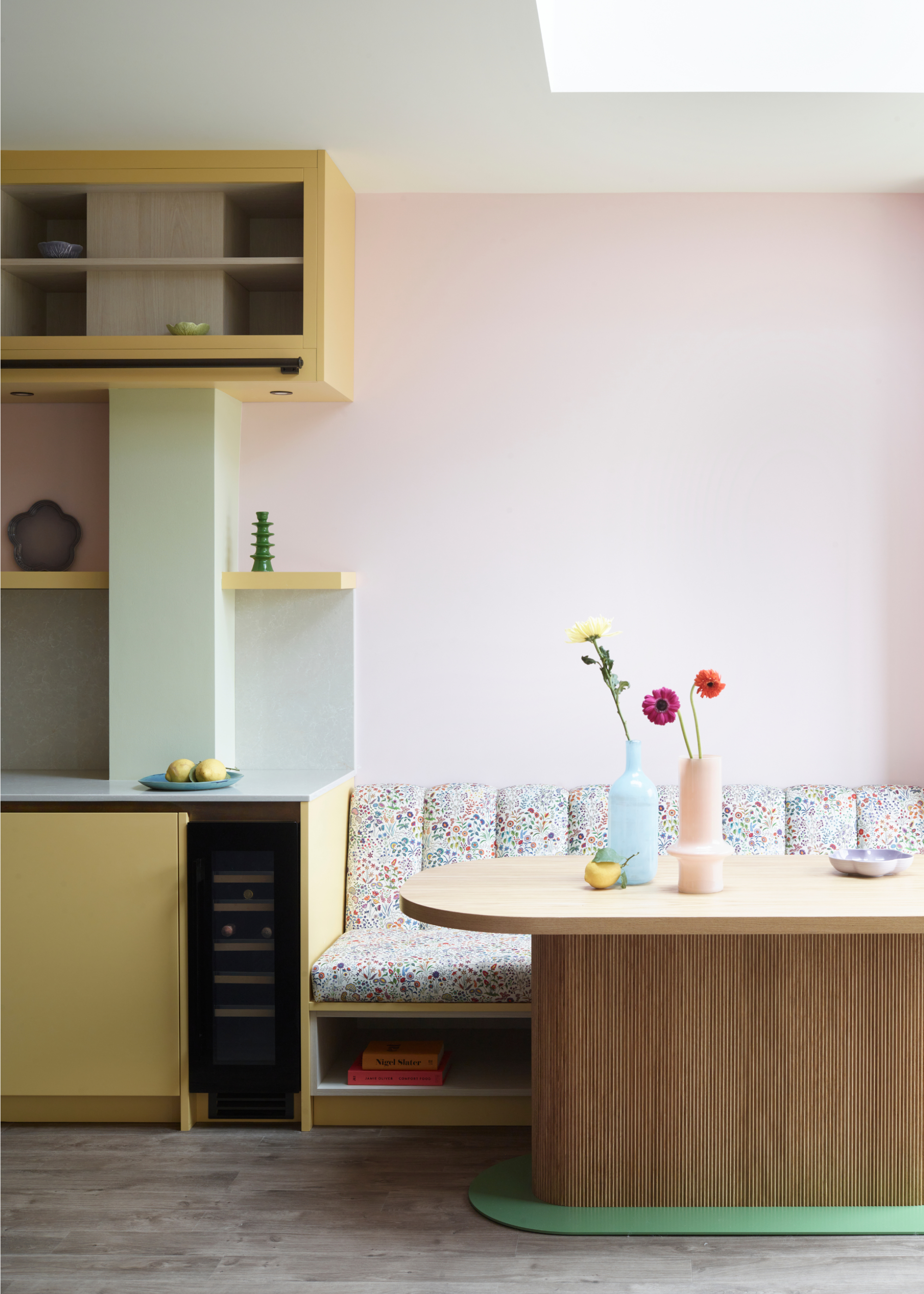 A modern style pastel kitchen finished in yellow cabinetry, green backsplash, and pink walls with patterned banquette seating behind a modern fluted oblong dining table