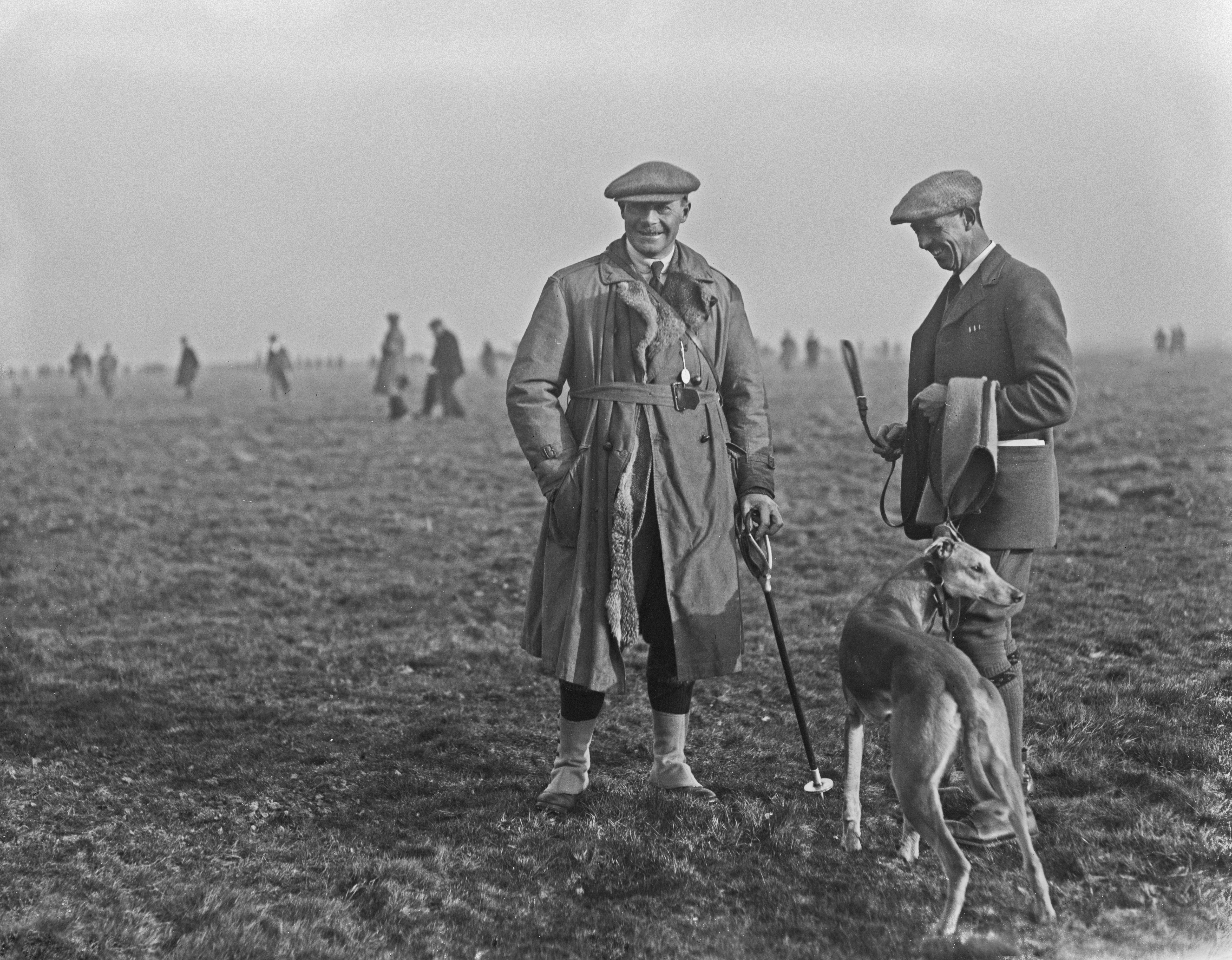 Two men dressed in early 20th-century country attire stand on a misty field, one holding a walking stick and the other holding a slender dog on a lead. More figures in coats and caps walk across the fog-covered landscape in the distance.