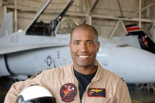 Astronaut Victor Glover stands in a flight suit in front of a jet, holding a helmet under his right arm.