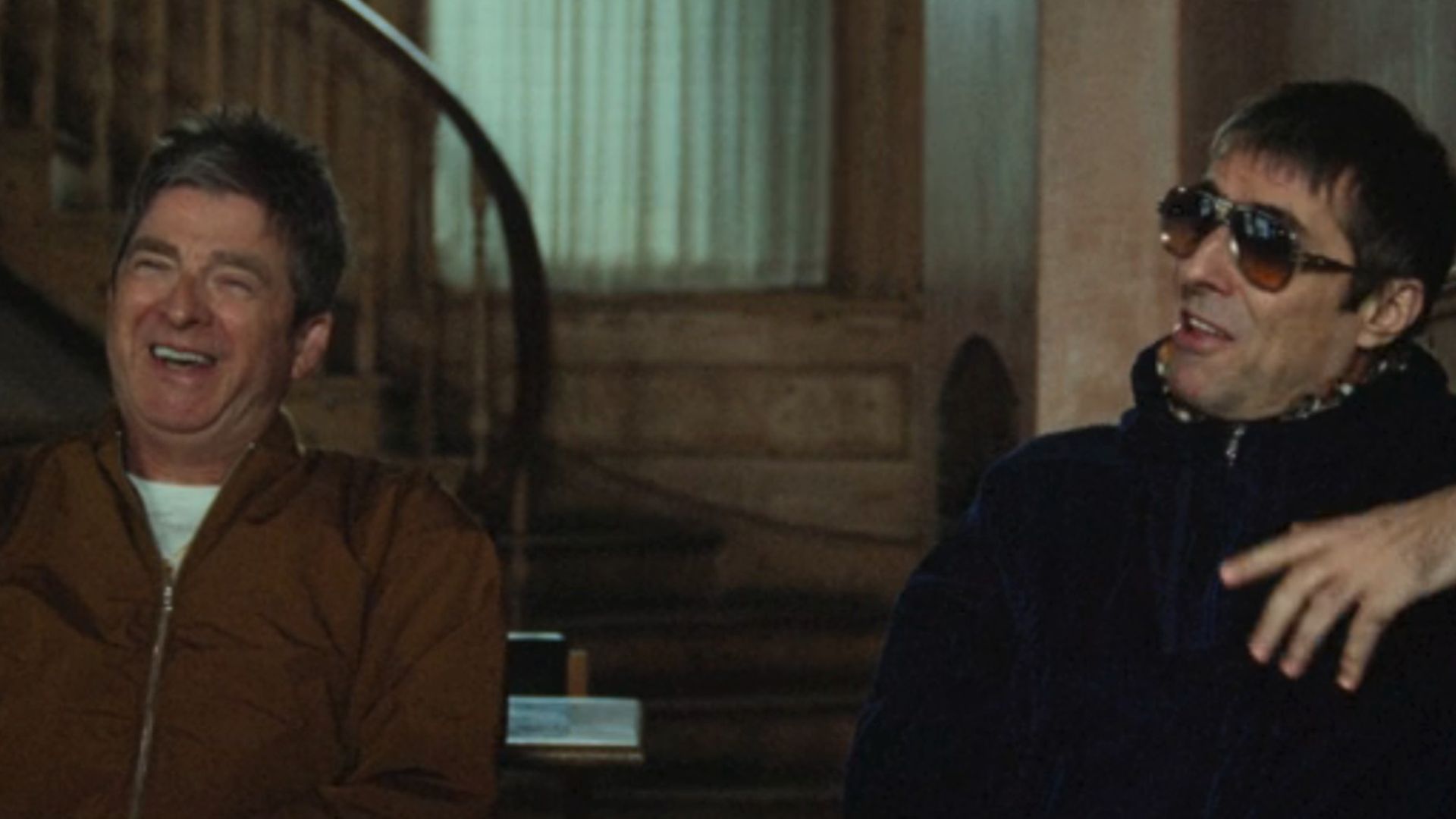 Noel Gallagher on the left, laughing, with Liam Gallagher on the right, wearing sunglasses and laughing in an interview together in Oasis: Don't Look Back In Anger