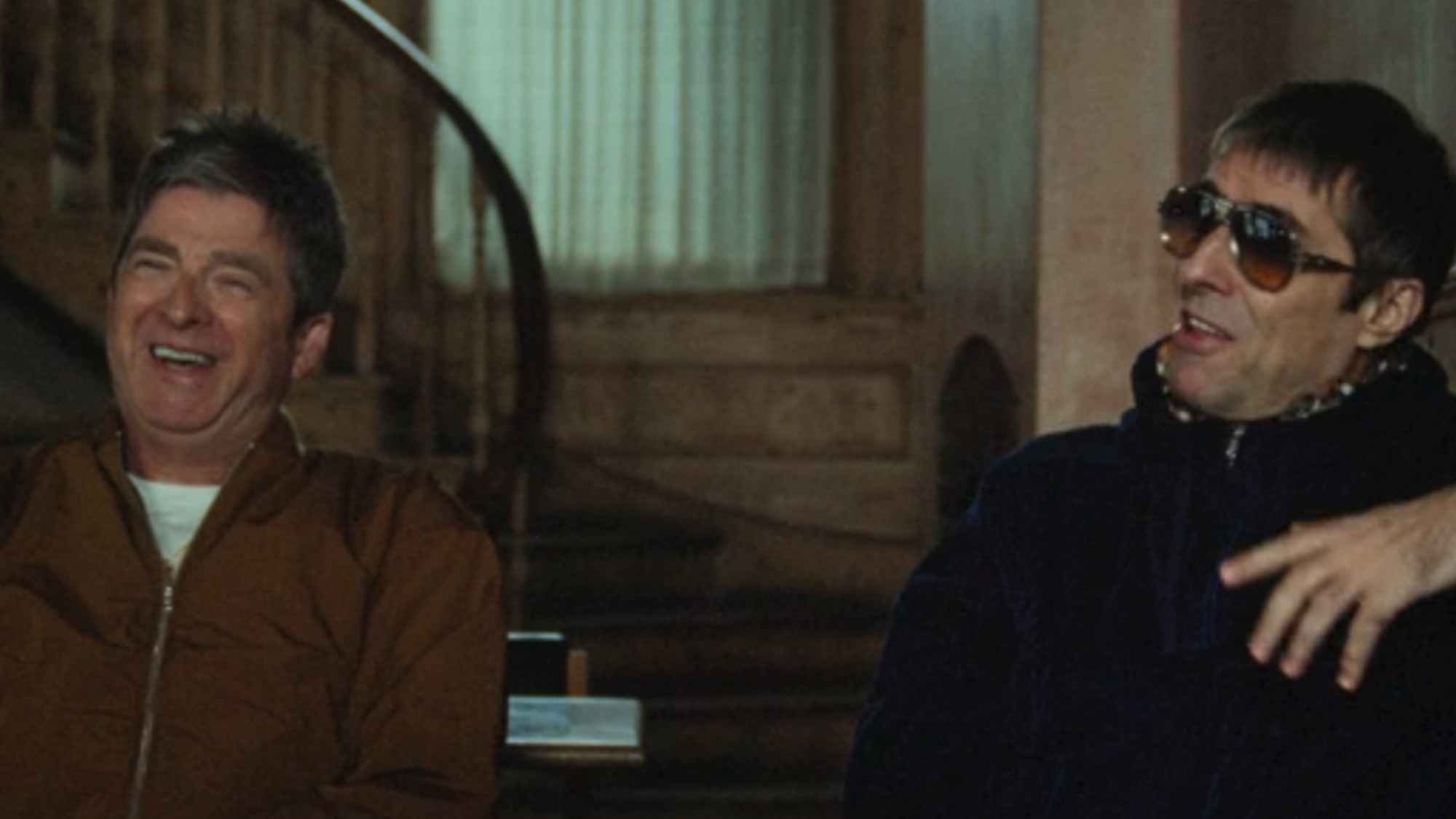 Noel Gallagher on the left, laughing, with Liam Gallagher on the right, wearing sunglasses and laughing in an interview together in Oasis: Don't Look Back In Anger