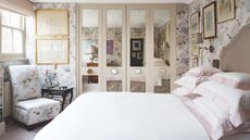 Small bedroom with mirrored four-door built-in closet, white bedding, small chair with side table and floral pastel wallpaper