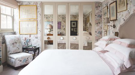Small bedroom with mirrored four-door built-in closet, white bedding, small chair with side table and floral pastel wallpaper