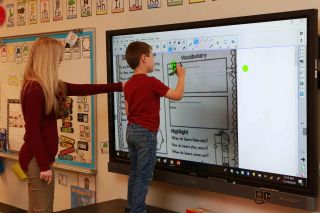 Teacher guides student in writing on BenQ flat panel display at front of class.