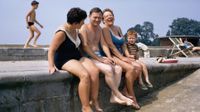 A candid, colorful 1960s photograph capturing a group of adults and a young boy laughing together while sitting with their legs dangling over a concrete sea wall at a public bathing area.