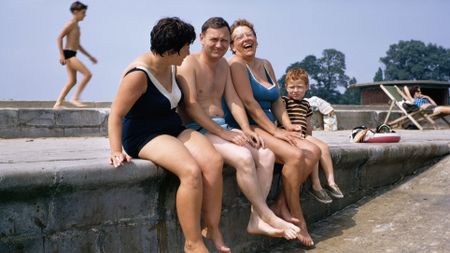 A candid, colorful 1960s photograph capturing a group of adults and a young boy laughing together while sitting with their legs dangling over a concrete sea wall at a public bathing area.