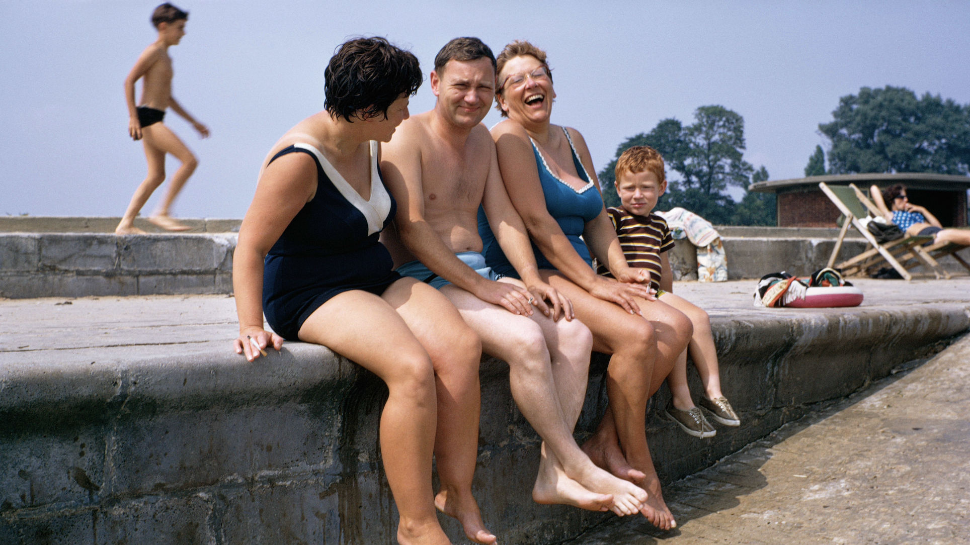 A candid, colorful 1960s photograph capturing a group of adults and a young boy laughing together while sitting with their legs dangling over a concrete sea wall at a public bathing area.