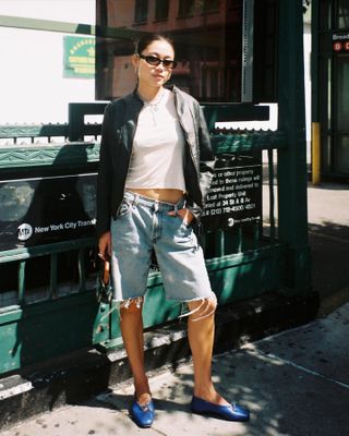 fashion influencer Vivian Li berpose di NYC dekat stasiun kereta bawah tanah mengenakan kacamata hitam bulat, jaket kulit tanpa kerah, kaus putih, celana pendek denim Bermuda, dan sepatu balet biru
