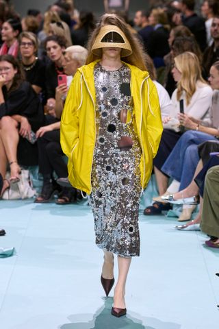 A model at the Prada S/S 25 show wearing a yellow nylon zip-up sporty jacket with a sequin midi dress, satin shoes, and a visor.