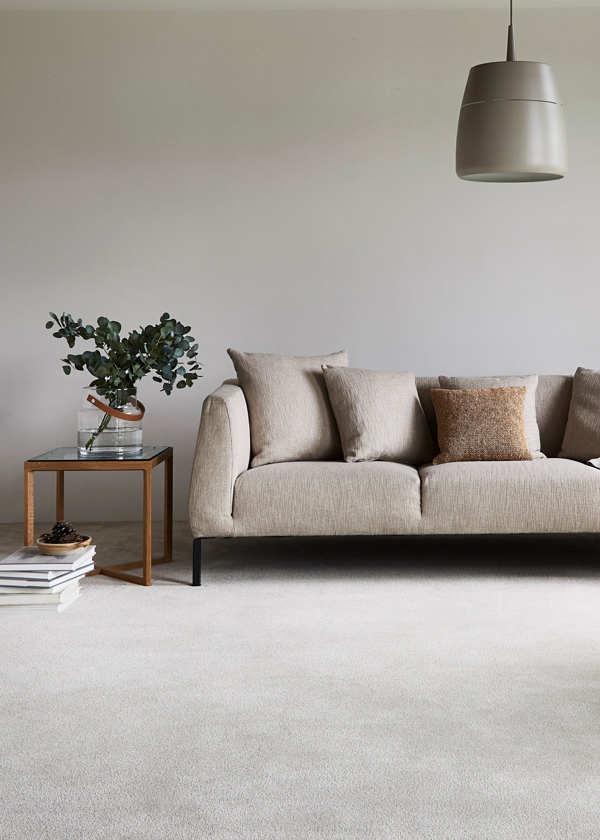 A nude colored sofa on a cream colored carpet. On the left side is a table with a vase and a plant inside.