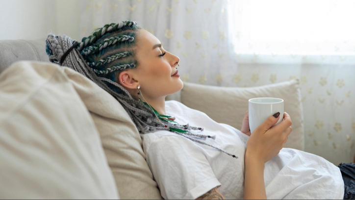 A woman with braided hair wearing a white t-shirt having a nap on sofa with a coffee cup in hand 
