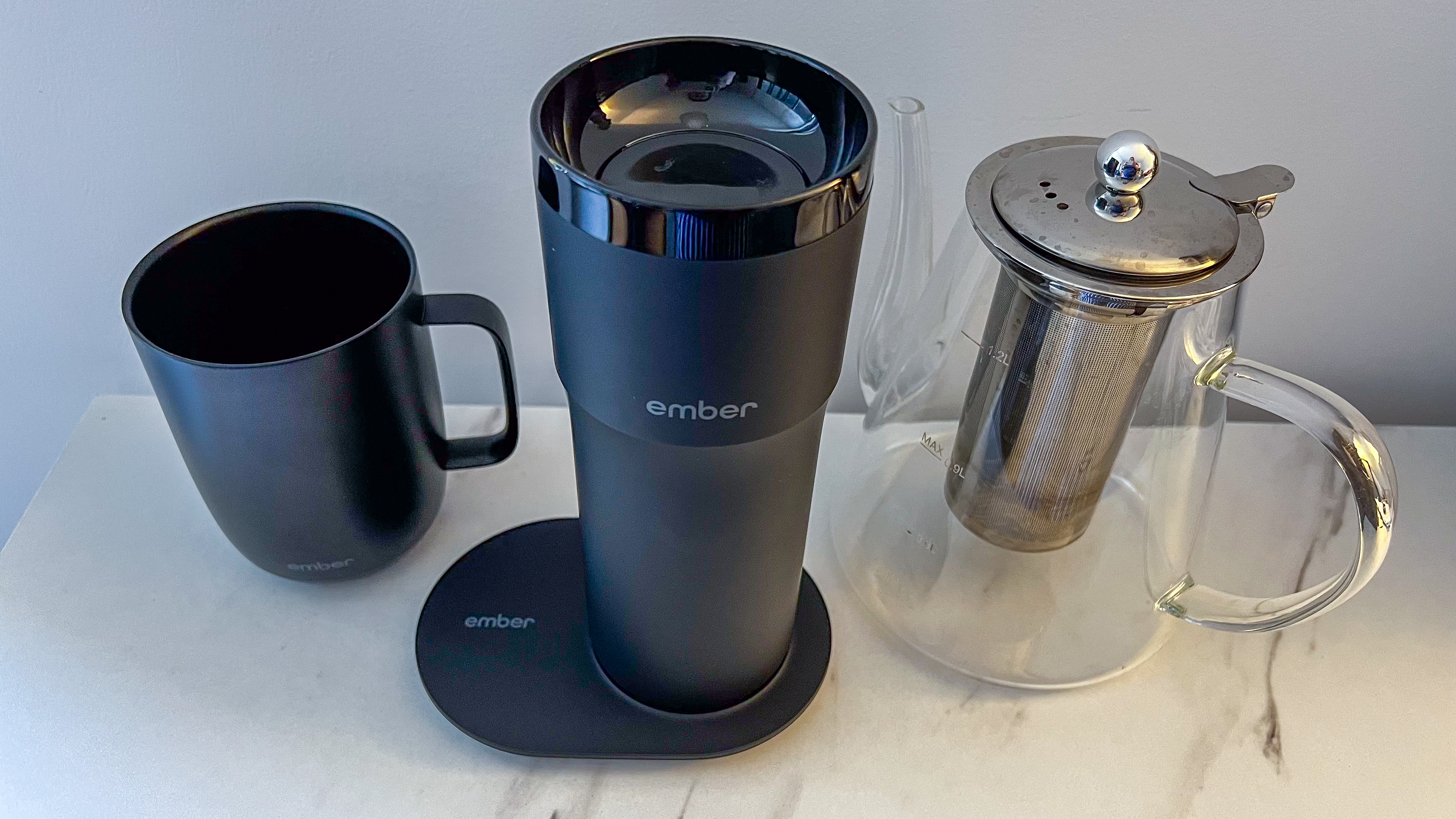 Ember Travel Mug 2+ on its charging plate, between Ember Mug 2 and a glass teapot