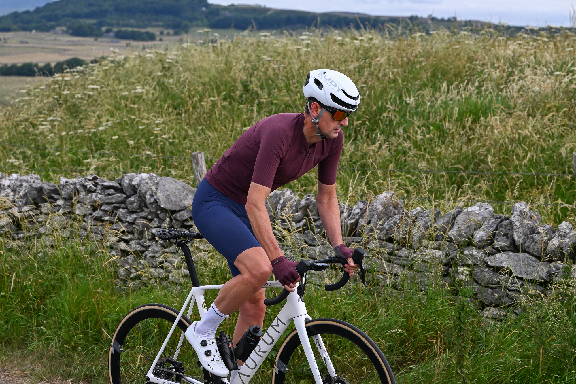 Man wearing a purple jersey, blue cycling shorts and a white helmet riding a white bike uphill out of the saddle