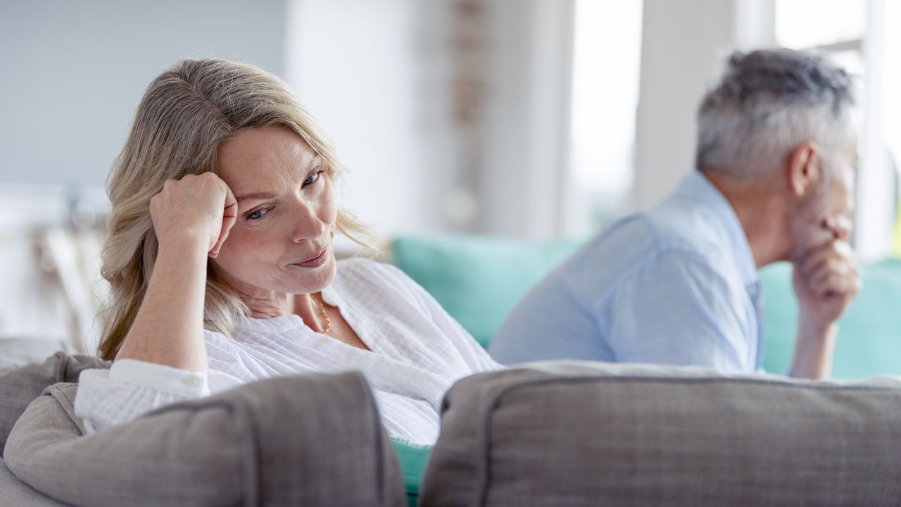 An older couple sit on the sofa looking sad and with their backs to each other.