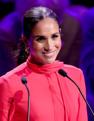 Meghan Markle in a red suit speaking behind a microphone