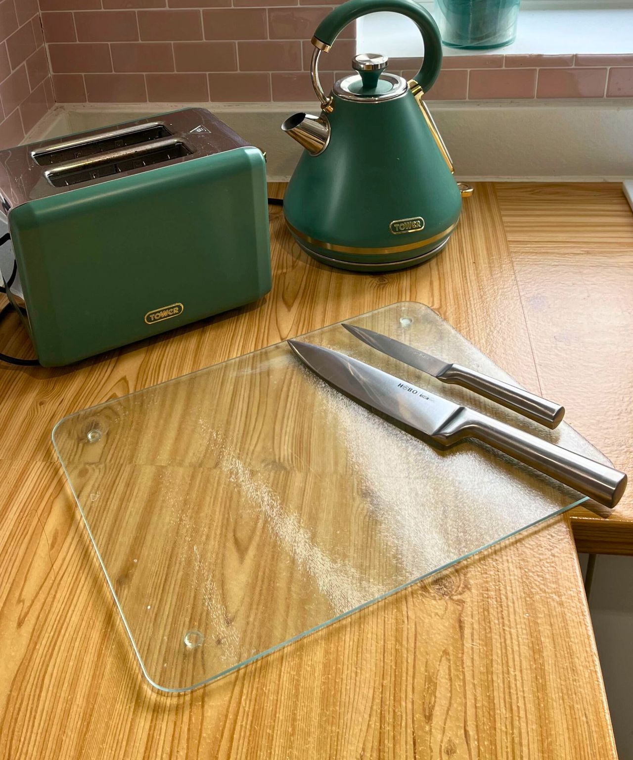Glass chopping board with two stainless steel knives on wooden countertop with green kettle and toaster in the background
