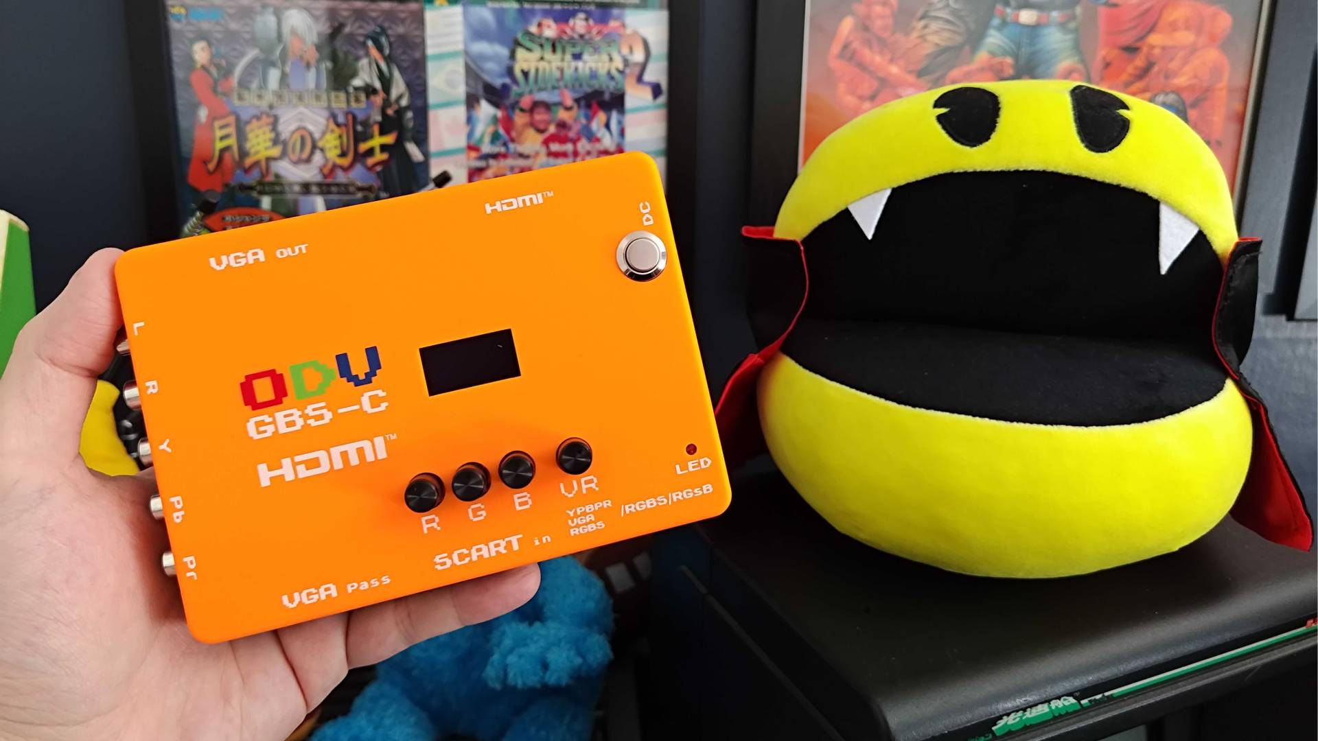 Hand holding ODV GBS-C upscaler with orange shell next to Vampire Pac-Man plush.