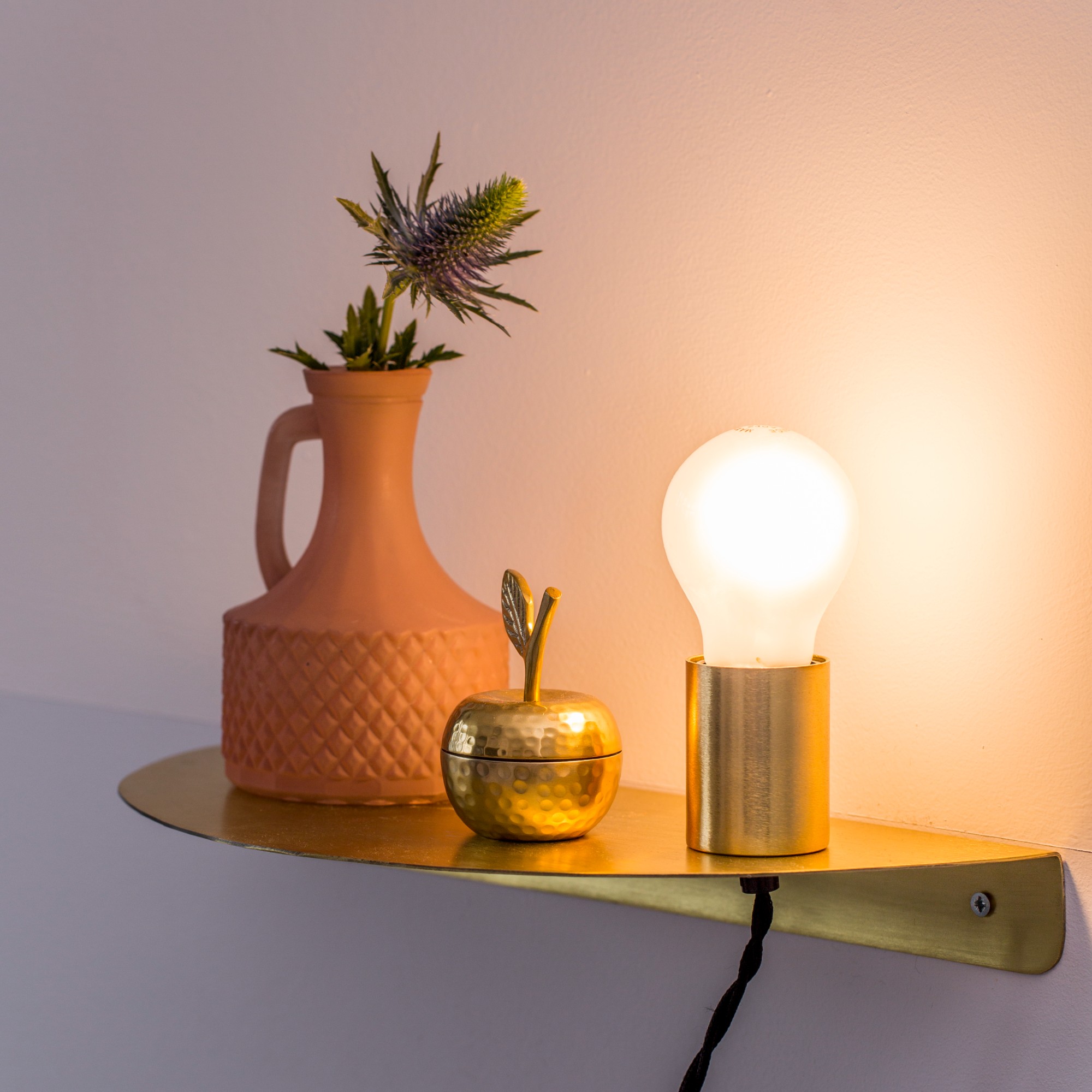 A brass curved shelf with a switched on table lamp with an exposed lightbulb in warm-tone light