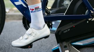 A white shoe and white sock are worn on a white leg on a bike on a turbo trainer