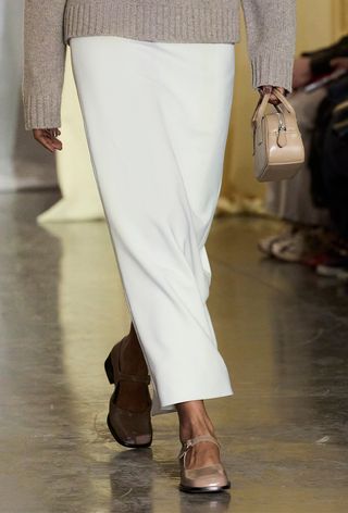 mary jane shoe trend is shown on Sandy Liangs's fall winter 2024 runway in a close photo of a tan mary janes paired with a white long pencil skirt, a tan sweater, and a tan bowler bag