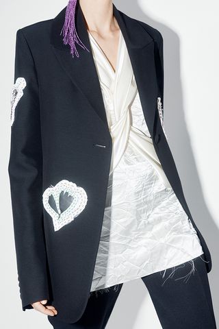 A torso view of a female model wearing black pants, a black jacket with white heart shapes on it and a white twisted shirt.