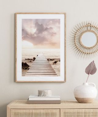 A brown wall with a sunset wall art print, a vase, a circiular mirroe, and a wooden console table with decor on it