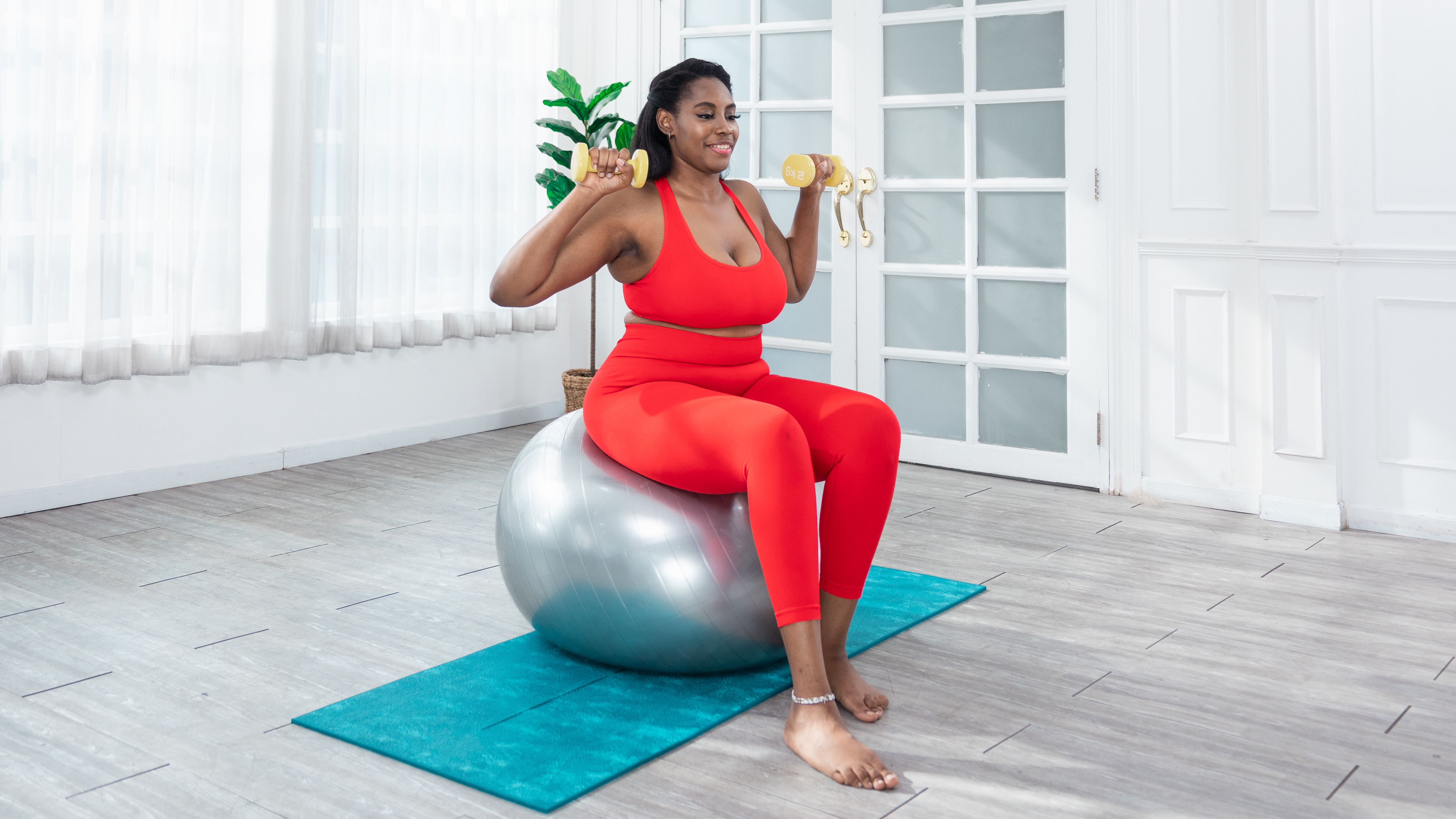 A woman sits on an exercise ball with her feet on the floor and light dumbbells held at her shoulders.