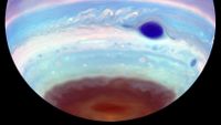 The southern hemisphere of Jupiter hangs from above in black space, striped in false color blues, violets, and dark reds