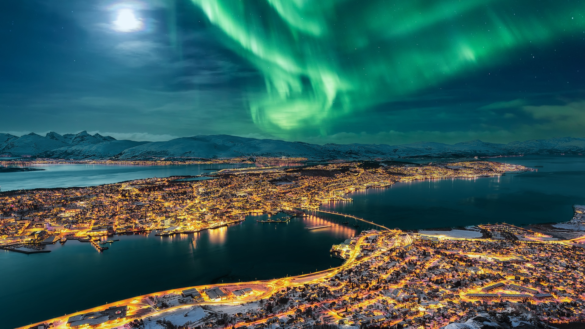 A cityscape illuminated by warm yellow lights sits below a stunning display of green northern lights sweeping across the night sky. Snow-covered mountains and a calm body of water add to the serene, wintery atmosphere.