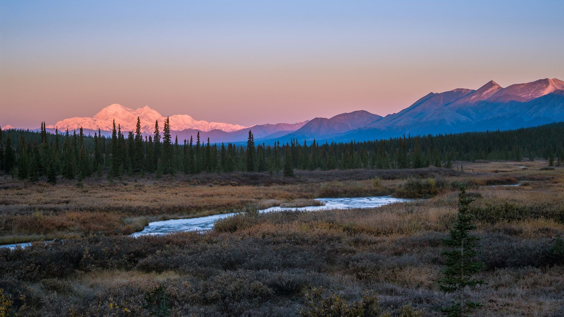 a lake in the foreground with trees in the background and mountains in the background, with a hint of pink.