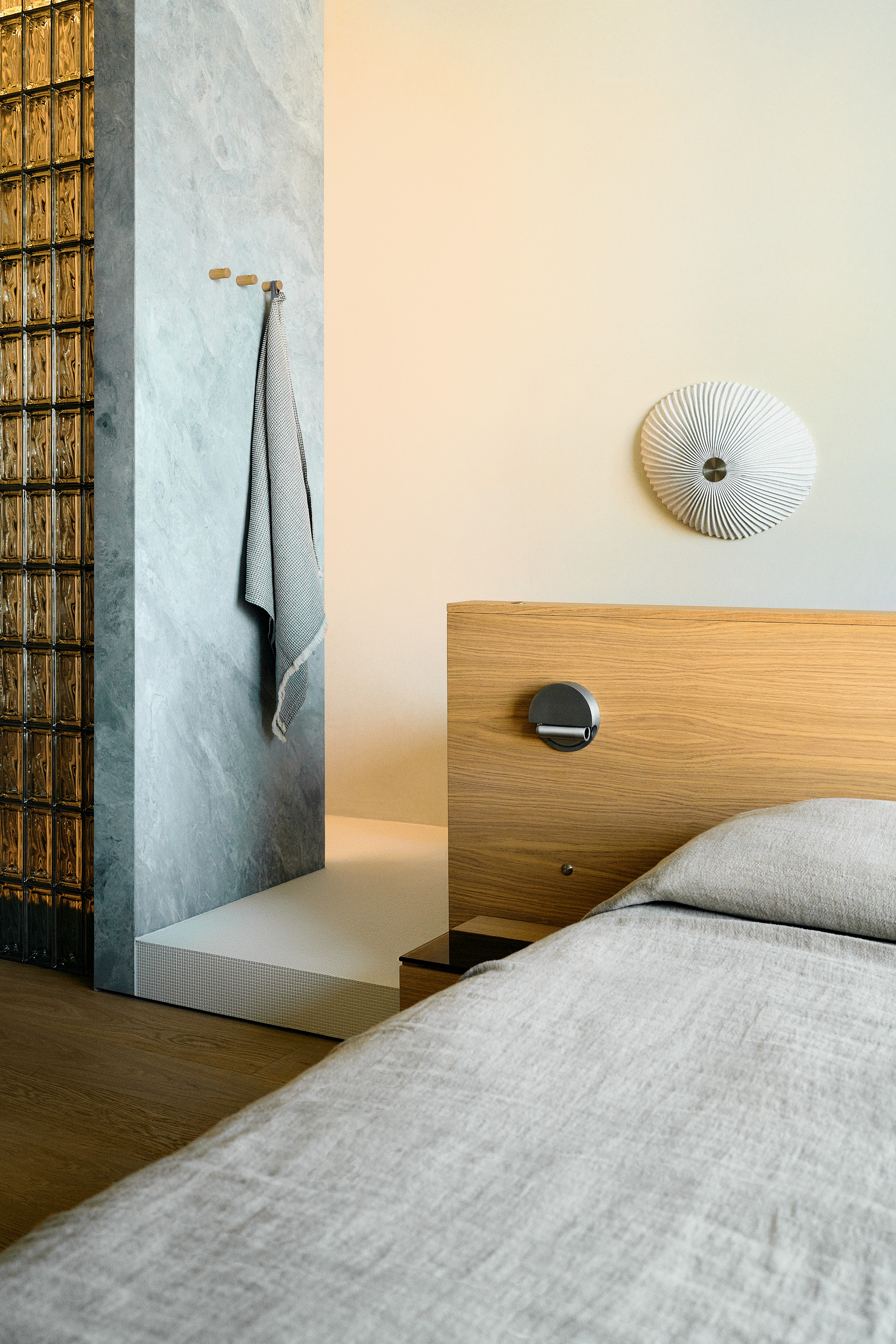 Bed with built-in headboard leading to white tiled platform housing shower area, with glass blocks and blue marble panel hiding it from view