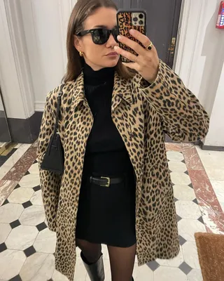 Die französische Mode-Influencerin Anne-Laure Mais posiert für ein Spiegel-Selfie und trägt einen Mantel mit Leopardenmuster, einen schwarzen Rollkragenpullover, einen schwarzen Minirock mit Gürtel, transparente schwarze Strumpfhosen und kniehohe schwarze Stiefel.