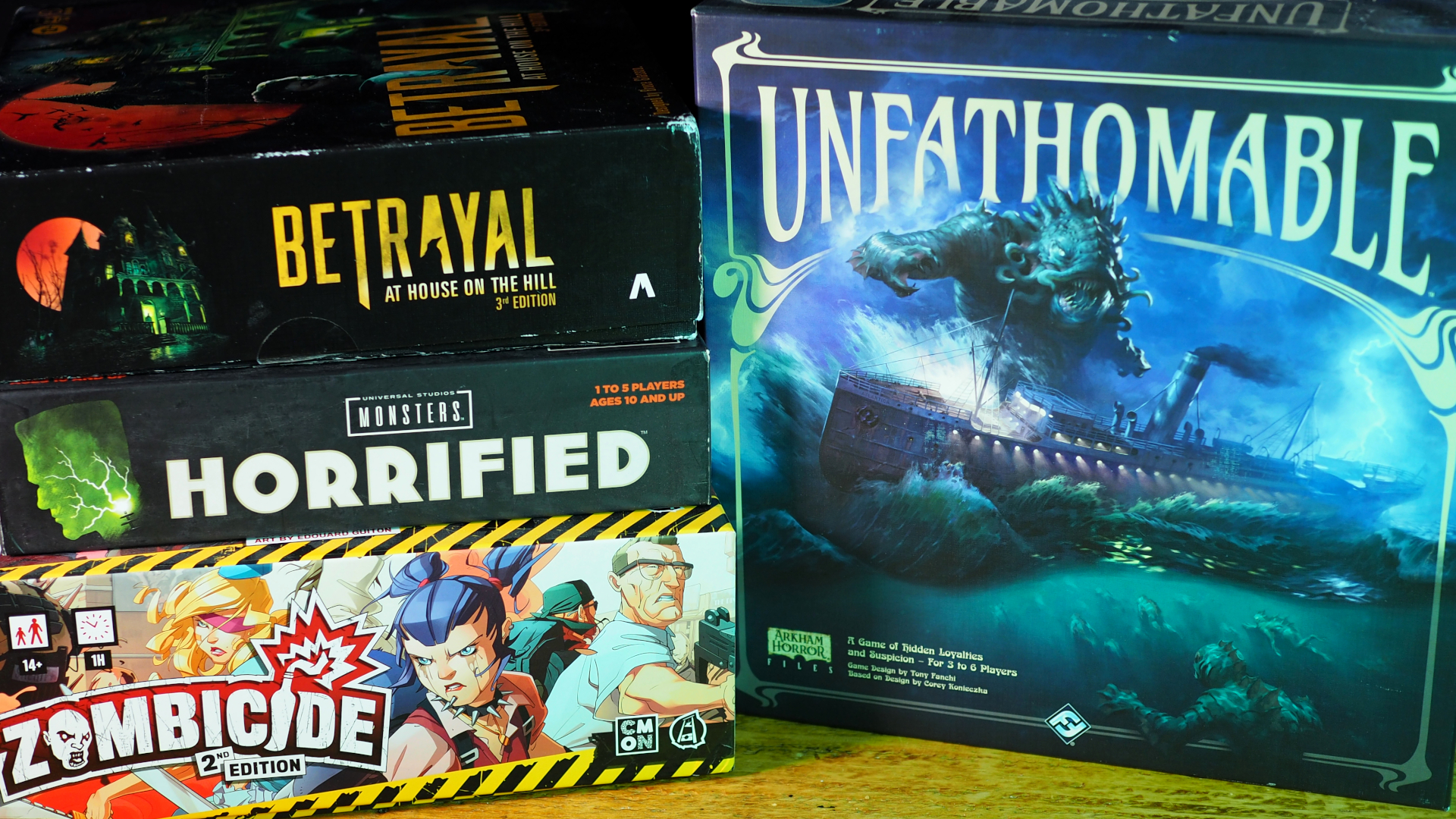 A collection of horror board games stacked beside each other, on a wooden table