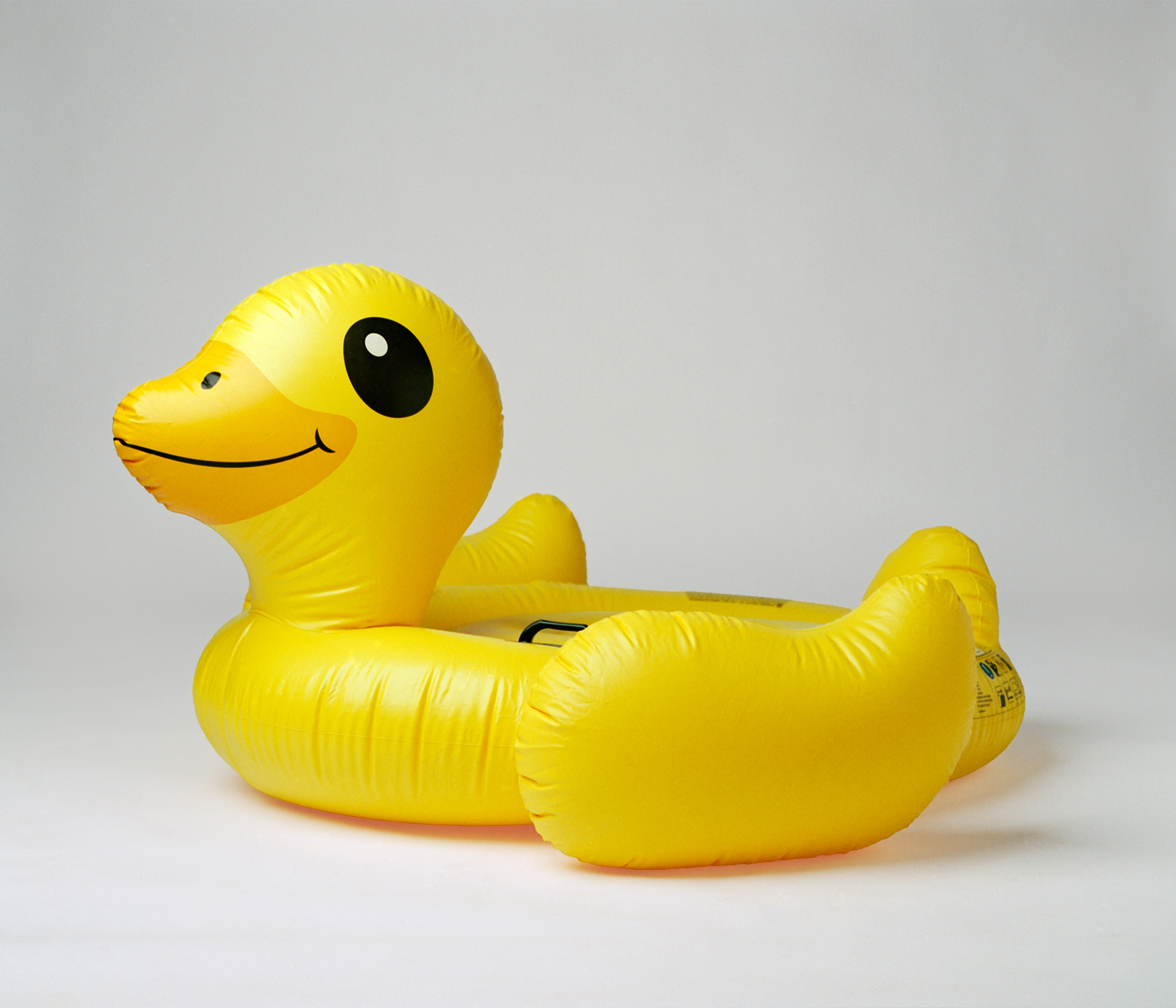 A bright yellow inflatable duck float against a plain white background. Its cheerful expression conveys a playful, summery vibe