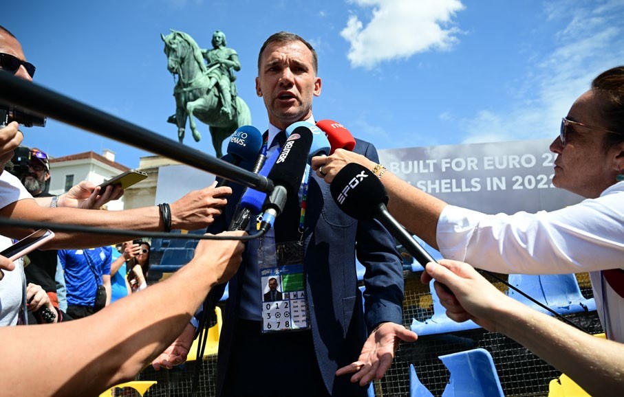 A suited football manager addresses the media surrounded by microphones, with a statue in the background, during a press event.