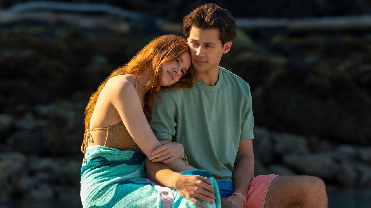 Percy and Sam sitting on a dock together, she's holding his arm and leaning into his sholder. 