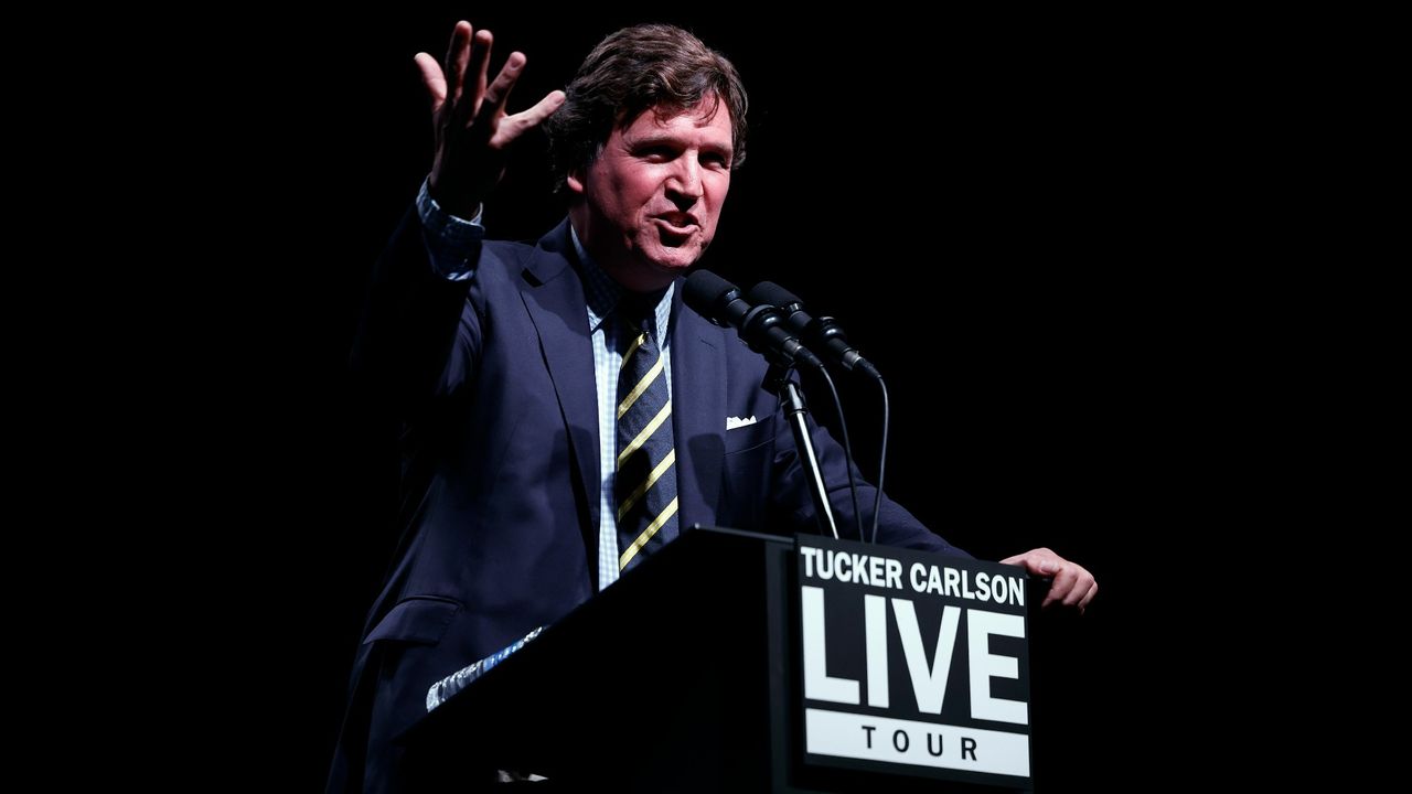 Tucker Carlson at a podium for a tour, dressed in a blue suit and a striped tie