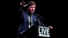 Tucker Carlson at a podium for a tour, dressed in a blue suit and a striped tie