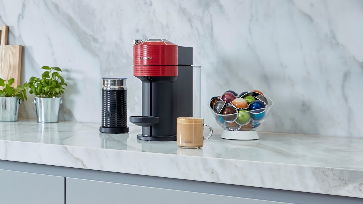 Die Nespresso Vertuo Next auf einer Arbeitsplatte, mit einem Kaffee und Kaffeekapseln daneben.