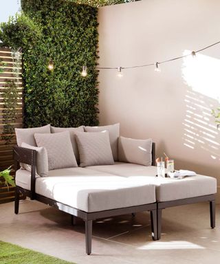 An outdoor day bed with a dark brown frame and light gray pillows and cushions on it with a grass wall to the back of it, a beige wall to the right of it, and black festoon lights strung above it