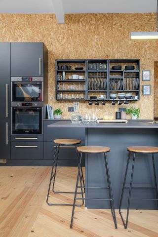 Black kitchen with open wall shelving, industrial bar stools, OSB wall and pine flooring