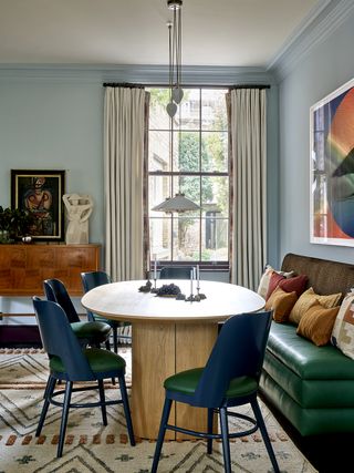 colorful dining room with blue walls, white ceiling, patterned rug, buffet in background with decor, a round timber dining table with four blue dining chairs and a green banquette seat and colorful cushions and a large artwork styled above it