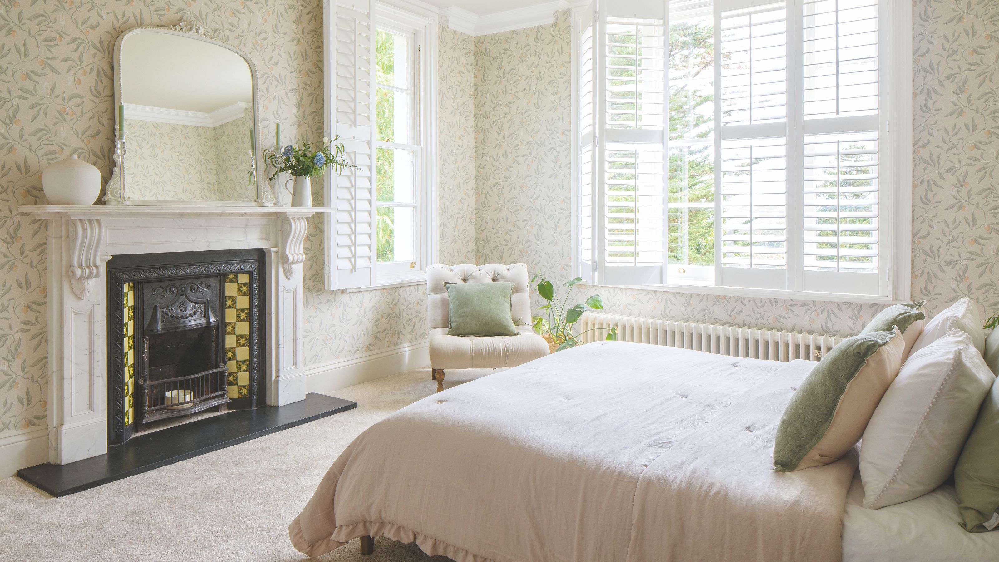 Cream bedroom with cream carpets, cream bedding, a fireplace in the middle, floral wallpaper and a cream accent chair